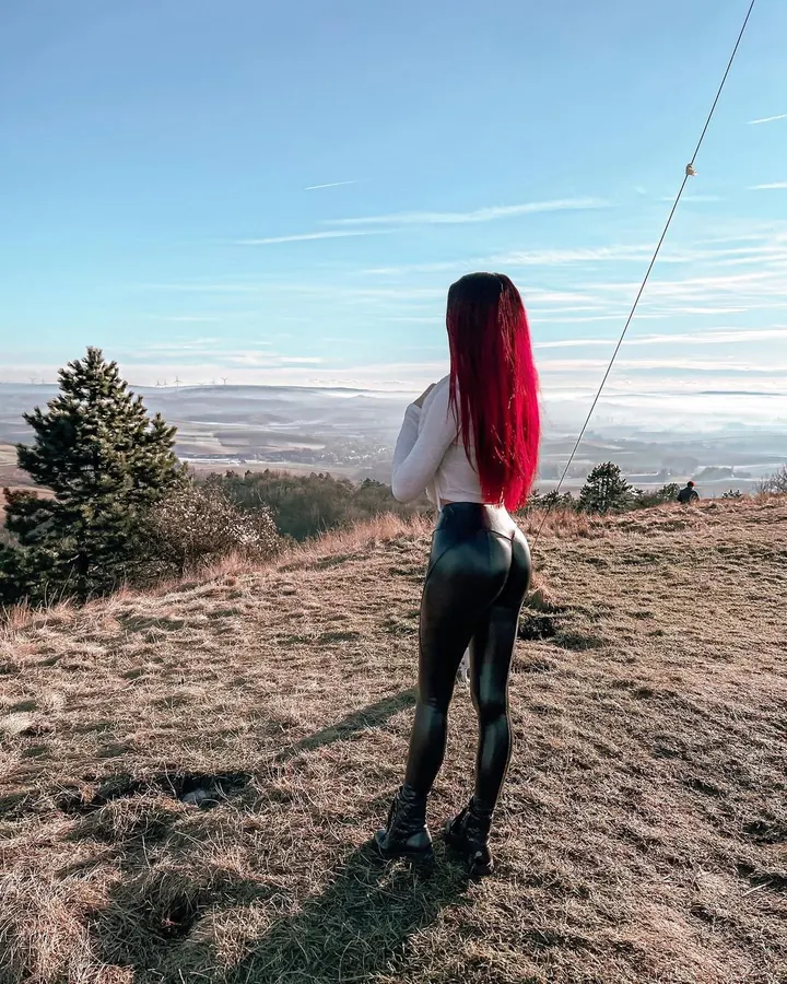Femme aux cheveux roux, peau blanche, en tenue grise ajustée, vue de dos devant un miroir, genoux pliés.