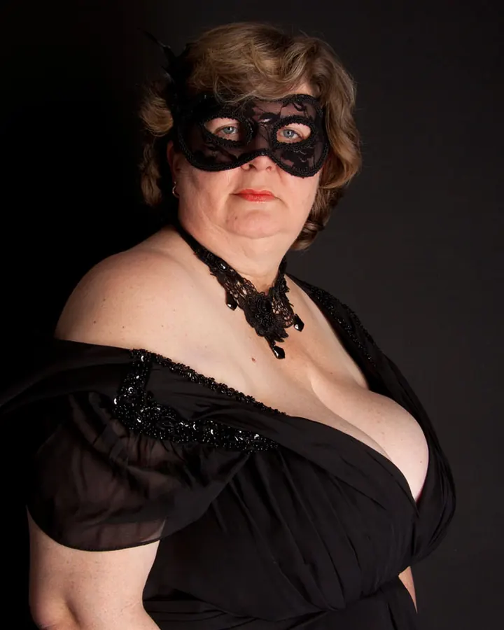 Femme blonde aux yeux marrons en robe noire, posant devant un fond noir, masque de dentelle sur les yeux