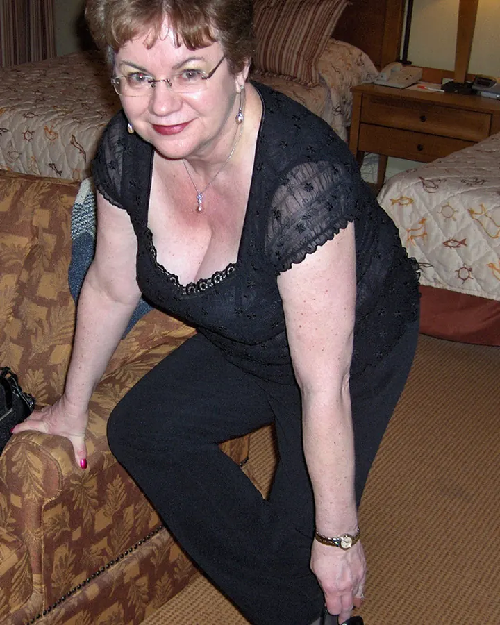 Femme blonde aux yeux bleus en robe noire, souriante, posant sur un canapé dans une chambre
