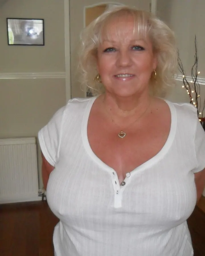 Femme blonde aux yeux bleus en haut blanc, souriante, posant dans un intérieur lumineux