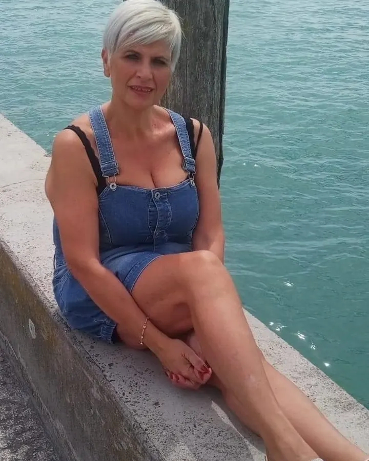 Femme aux cheveux blancs courts en salopette bleue, assise sur un mur de béton près de l'eau.