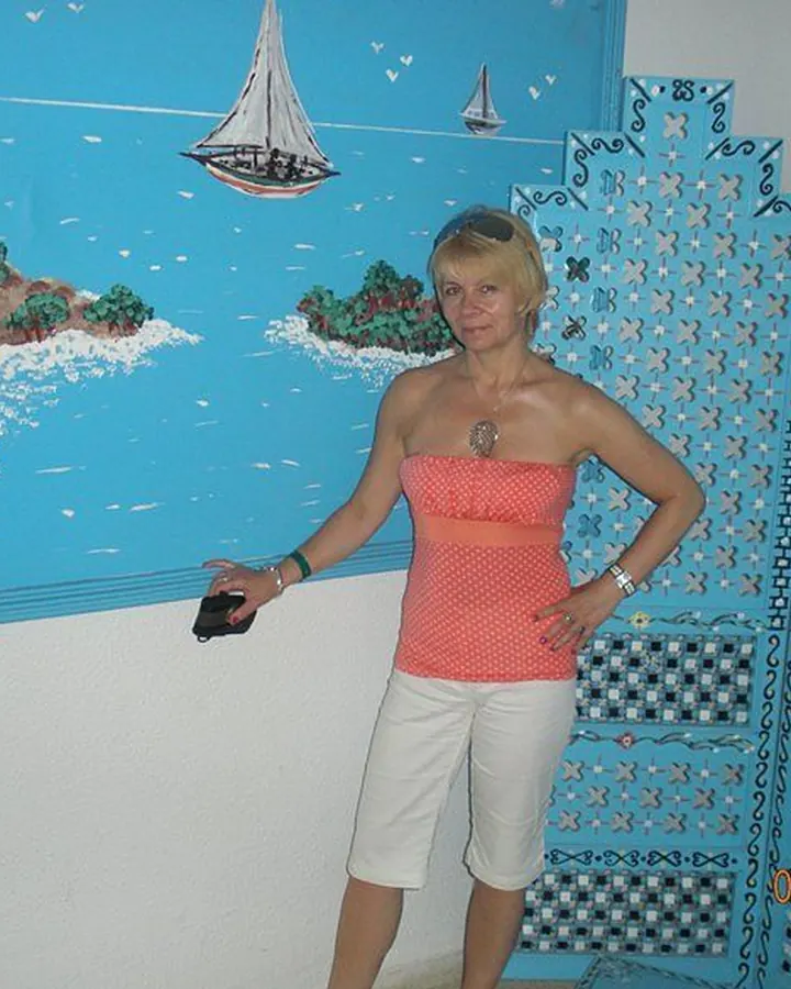 Femme aux cheveux blonds en haut rose à pois et pantalon blanc, debout devant un mur bleu avec une peinture marine.