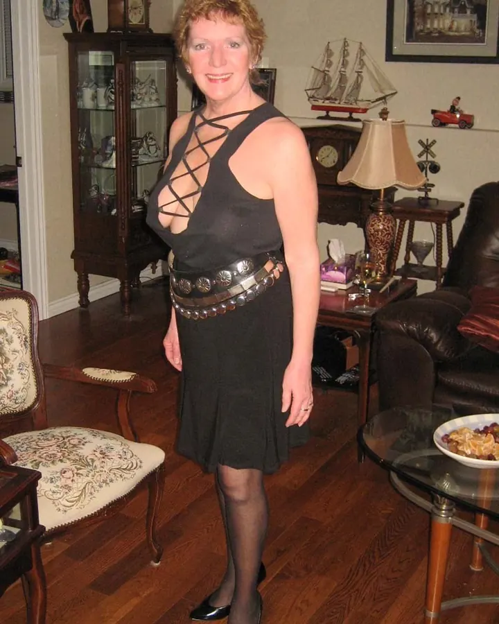 Femme aux cheveux roux, en robe noire avec lacets, ceinture décorative, collant noir, chaussures noires, dans un salon classique.
