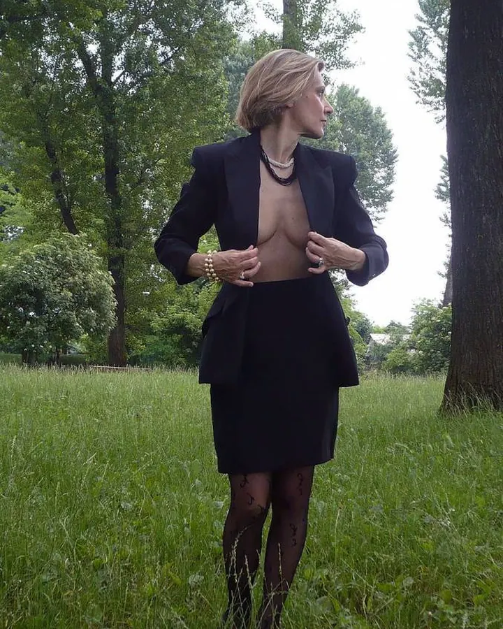 Femme aux cheveux blonds, en robe noire, collant noir, pose sur un lit, main sur la hanche, regard direct.