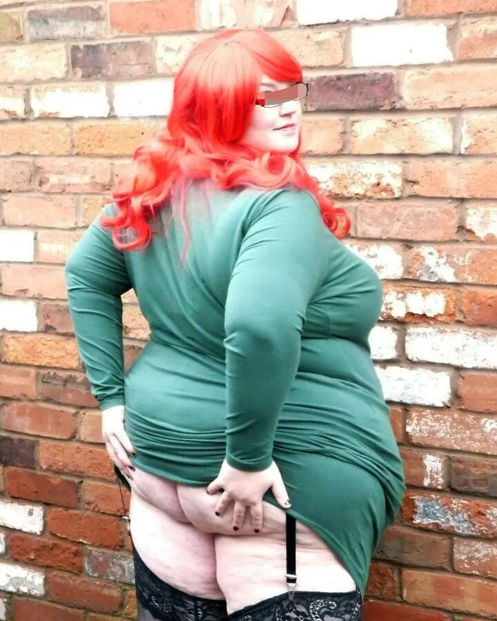 Femme aux cheveux roux portant une robe verte et des bas noirs, debout devant un mur de briques.