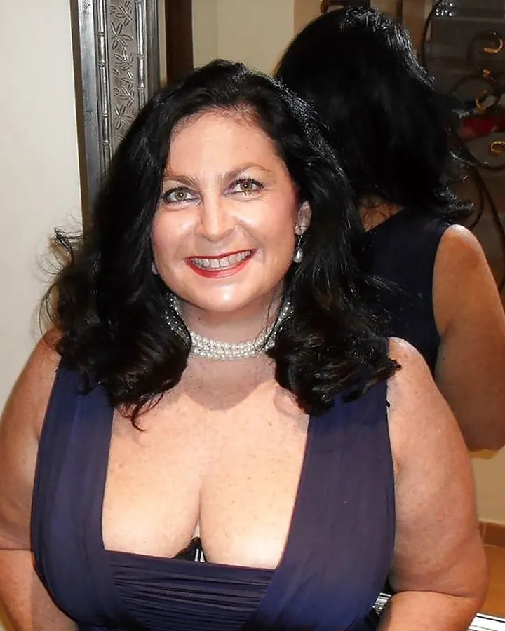 Femme aux cheveux noirs, portant une robe noire à motifs floraux, souriant à la caméra dans un intérieur.