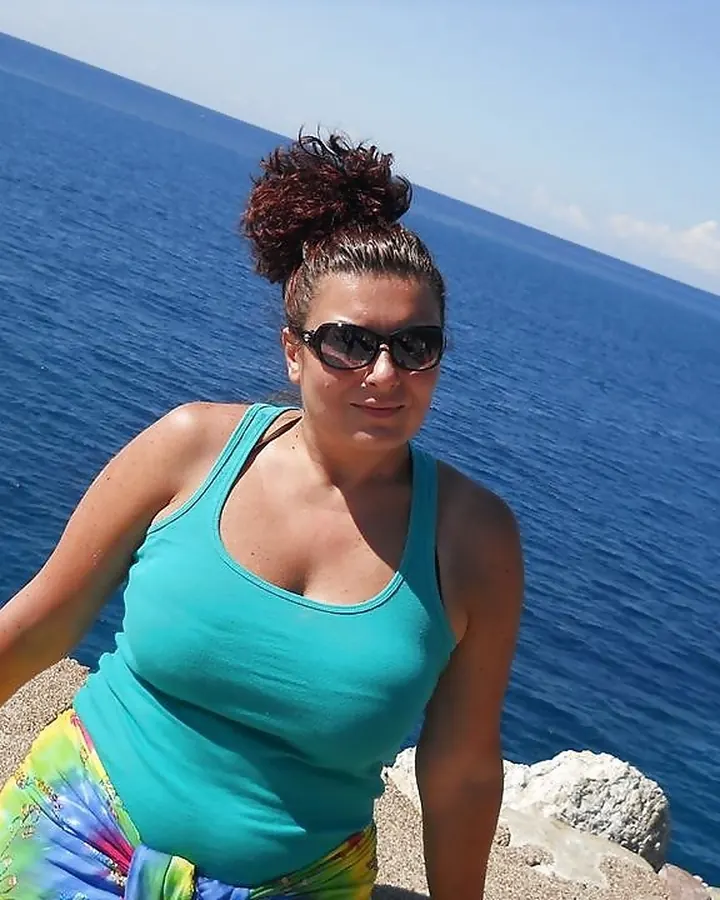 Femme aux cheveux bruns, portant un bikini rose à motifs serpent, une jupe colorée et des bijoux, posant sur un bateau en mer.