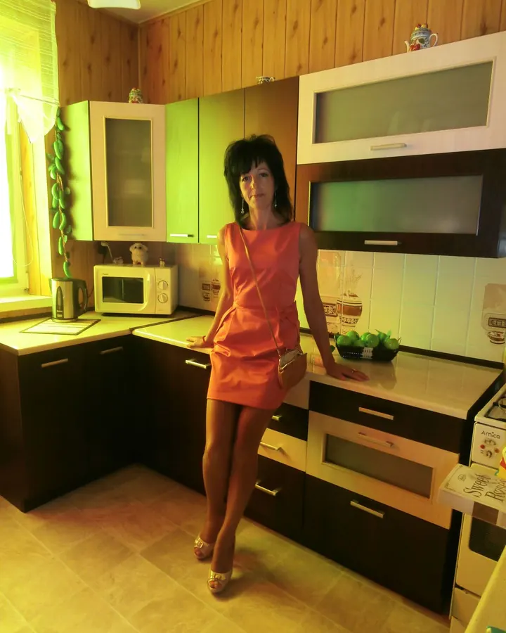 Femme aux cheveux bruns-noirs portant une robe orange, debout dans une cuisine, les mains sur les plans de travail, avec un four, un micro-ondes et des fruits en arrière-plan.