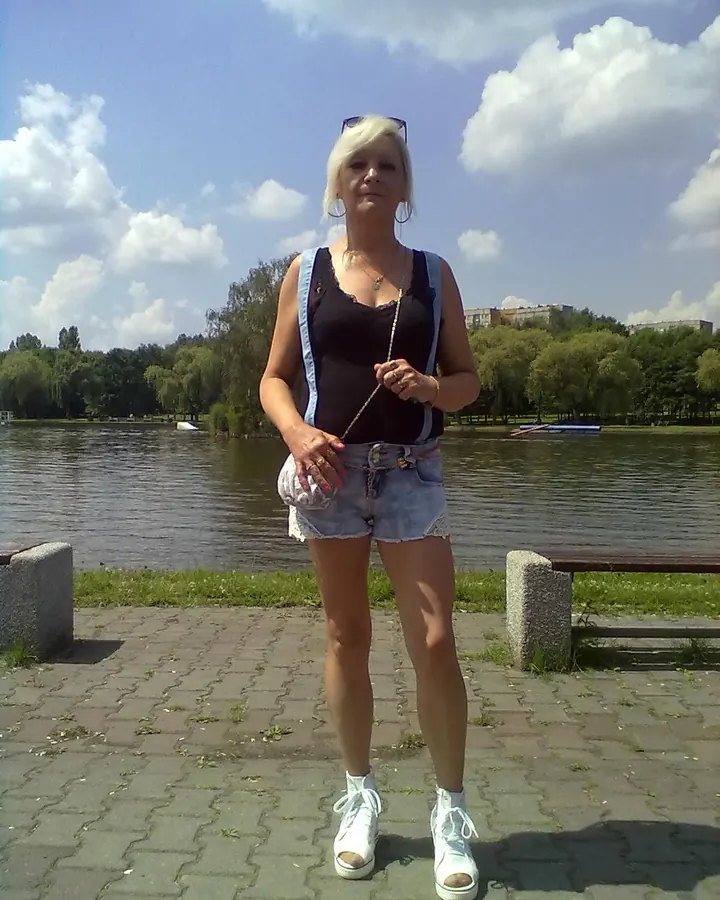 Femme blonde portant un haut noir et un short en jean délavé, debout près d'un lac, les mains sur les hanches, avec lunettes de soleil sur la tête, sous un ciel bleu nuageux.