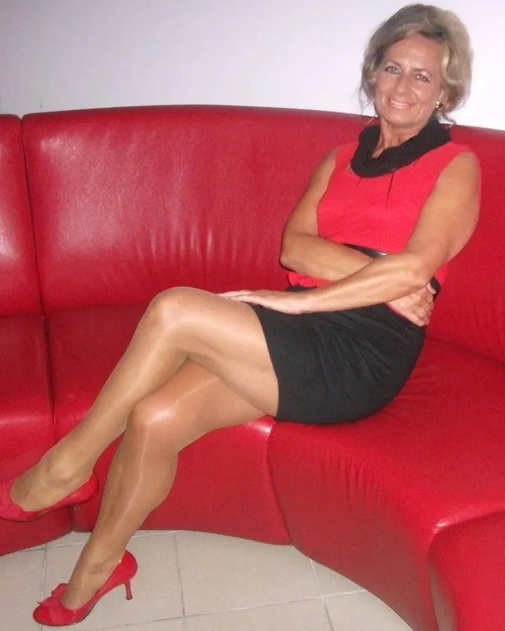 Femme aux cheveux blonds courts, souriante, portant un haut noir et une jupe rouge, assise sur un canapé rouge.