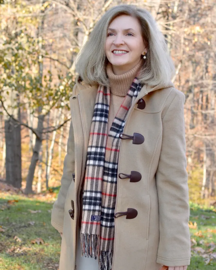 Femme aux cheveux gris-argentés, souriante, portant un manteau beige, un pull beige, une écharpe à carreaux et une pochette marron, debout dans un parc automnal.