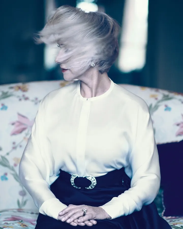 Femme aux cheveux blonds courts, souriante, portant un haut noir et une jupe rouge, assise sur un canapé rouge.