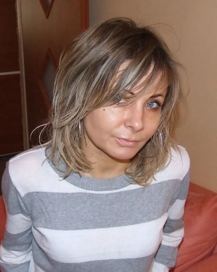 Femme aux cheveux blonds portant un body noir, assise sur un lit blanc, les mains sur les hanches.