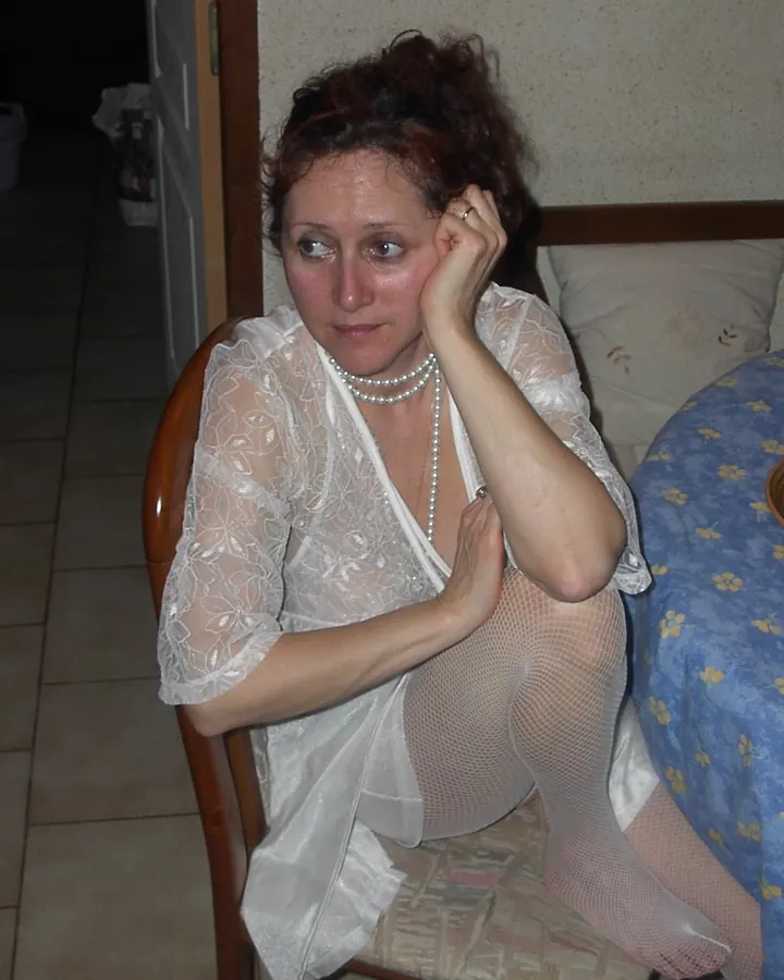 Femme aux cheveux châtains portant un haut blanc et un short en jean, debout devant un armoire blanche dans une chambre rose.