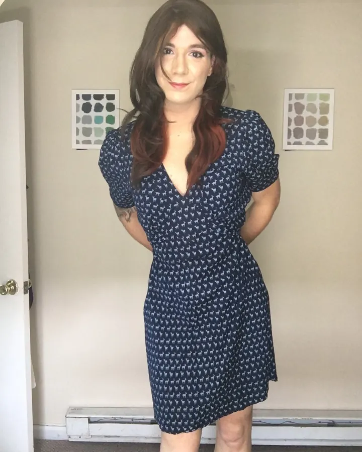 Femme en robe bleu marine à motifs, mains derrière le dos, posant dans un couloir.