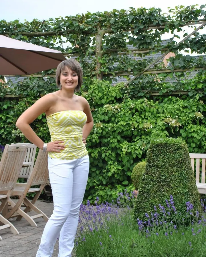 Femme en haut jaune à motifs, pantalon blanc, posant dans un jardin verdoyant avec des fleurs de lavande.