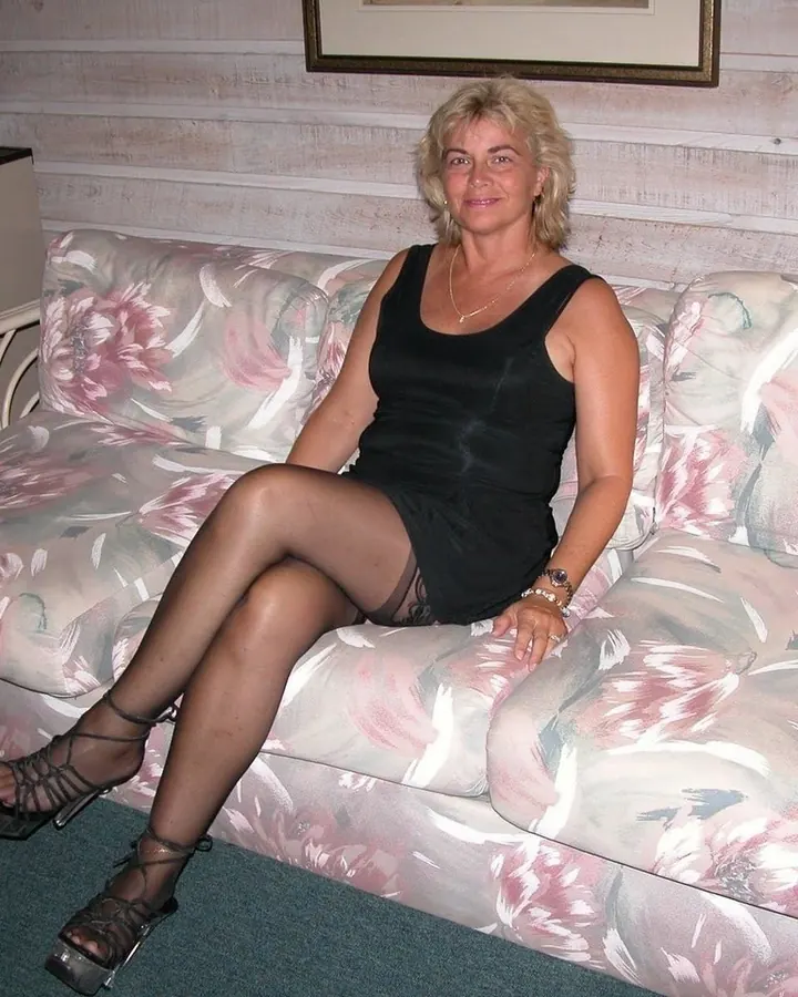 Femme aux cheveux blonds portant une robe noire, assise sur un canapé à motifs floraux, les jambes croisées