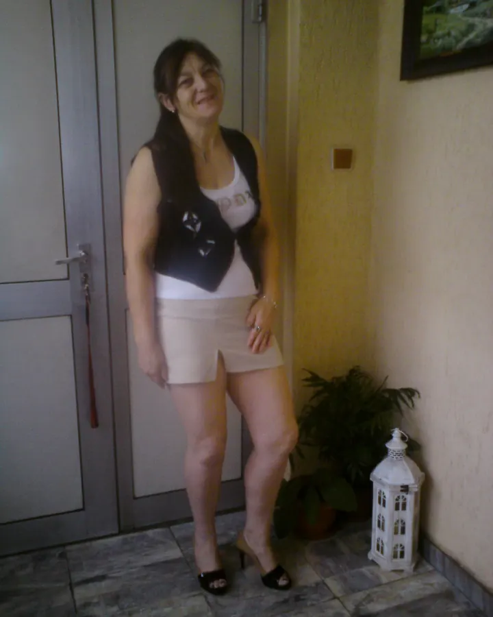 Femme aux cheveux châtains longs, souriante, en haut blanc, gilet noir et jupe beige, debout près d'une porte intérieure.